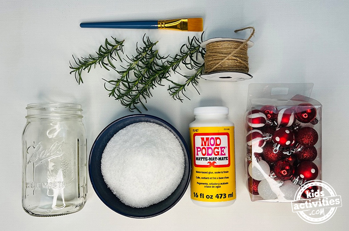 Frosted candle Christmas craft-Image shows the supplies needed to make a christmas craft: mason ja, epsom salt, mod podge, ornaments, paintbrush, twine, twigs. from Kids Activities Blog