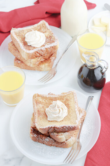 Easy Eggnog French Toast - Two white plates with Eggnog French Toast topped with powdered sugar and whipped cream, glasses of orange juice, and a glass container of maple syrup - Kids Activities Blog