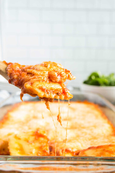 A wooden spoon holding two ravioli with a pan of cheesy baked ravioli in the background, along with a green salad.