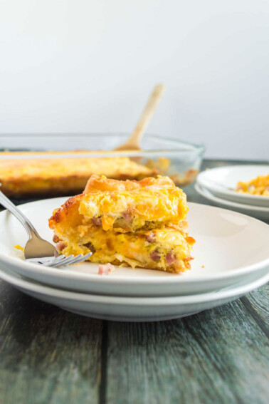 A piece of ham and egg breakfast casserole sitting on a white plate with a 9x13 pan of casserole in the background, along with some more white plates.