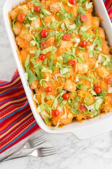 A white 9x13 pan containing tater tot taco casserole, garnished with fresh shredded lettuce and halved cherry tomatoes.