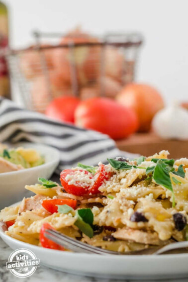 White Plate with Greek Pasta Salad on it with a basket of onions and tomatoes in the background