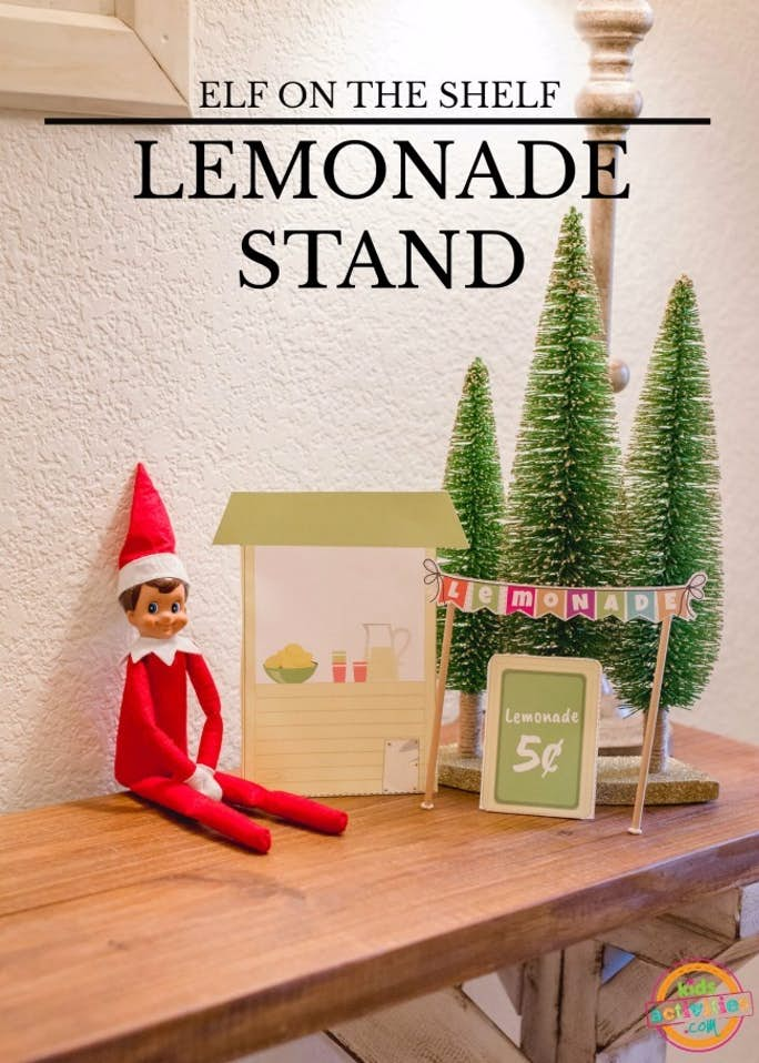 Text: Elf on the Shelf Lemonade Stand- Kids Activities Blog- Elf on the Shelf sitting against a w all with the lemonade stand and a sign that says lemonade 5 cents and a lemonade banner with Christmas trees in the background